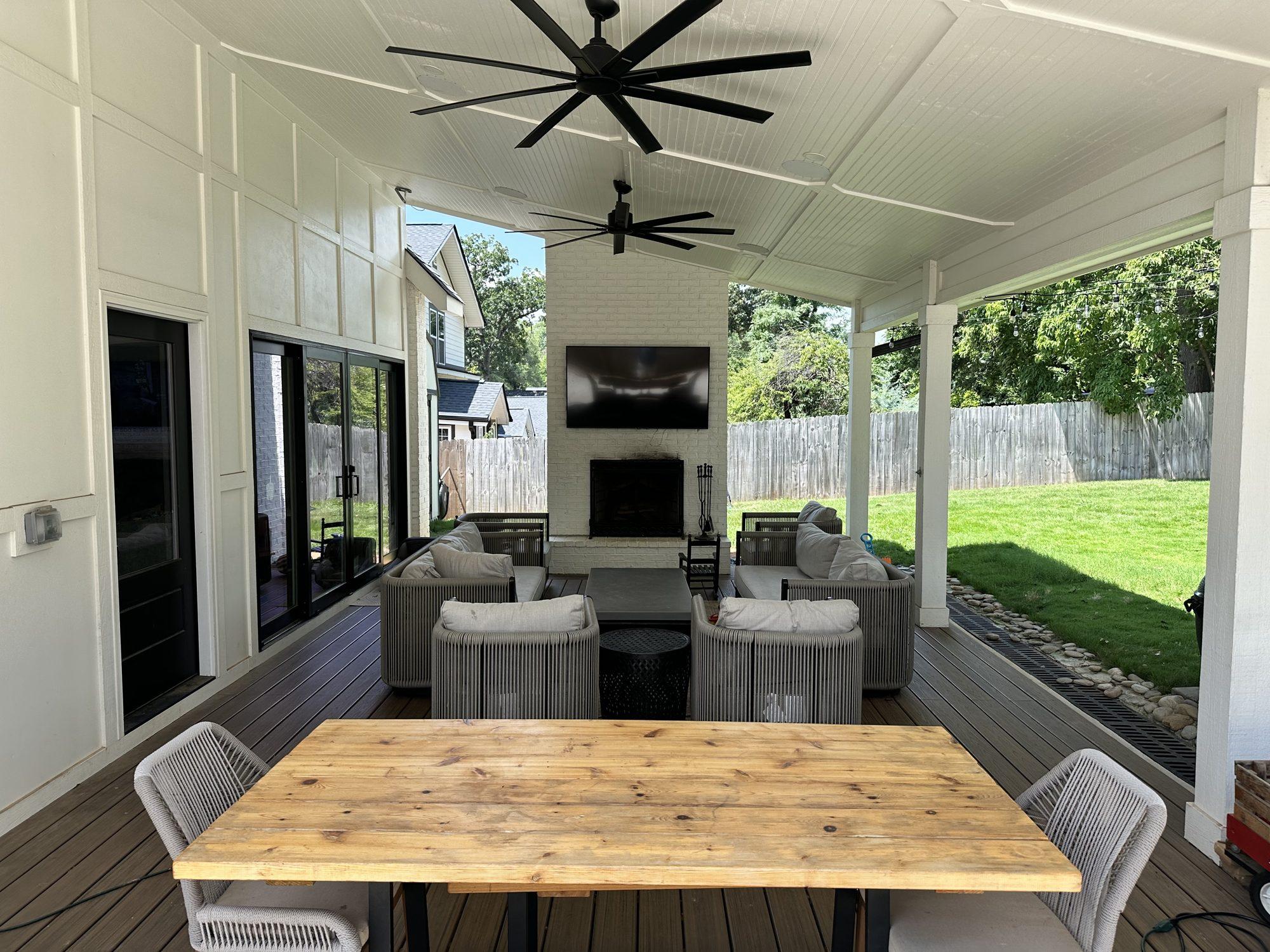 Covered outdoor living space with a painted brick fireplace, mounted TV, ceiling fans, and a wood dining table overlooking the yard.